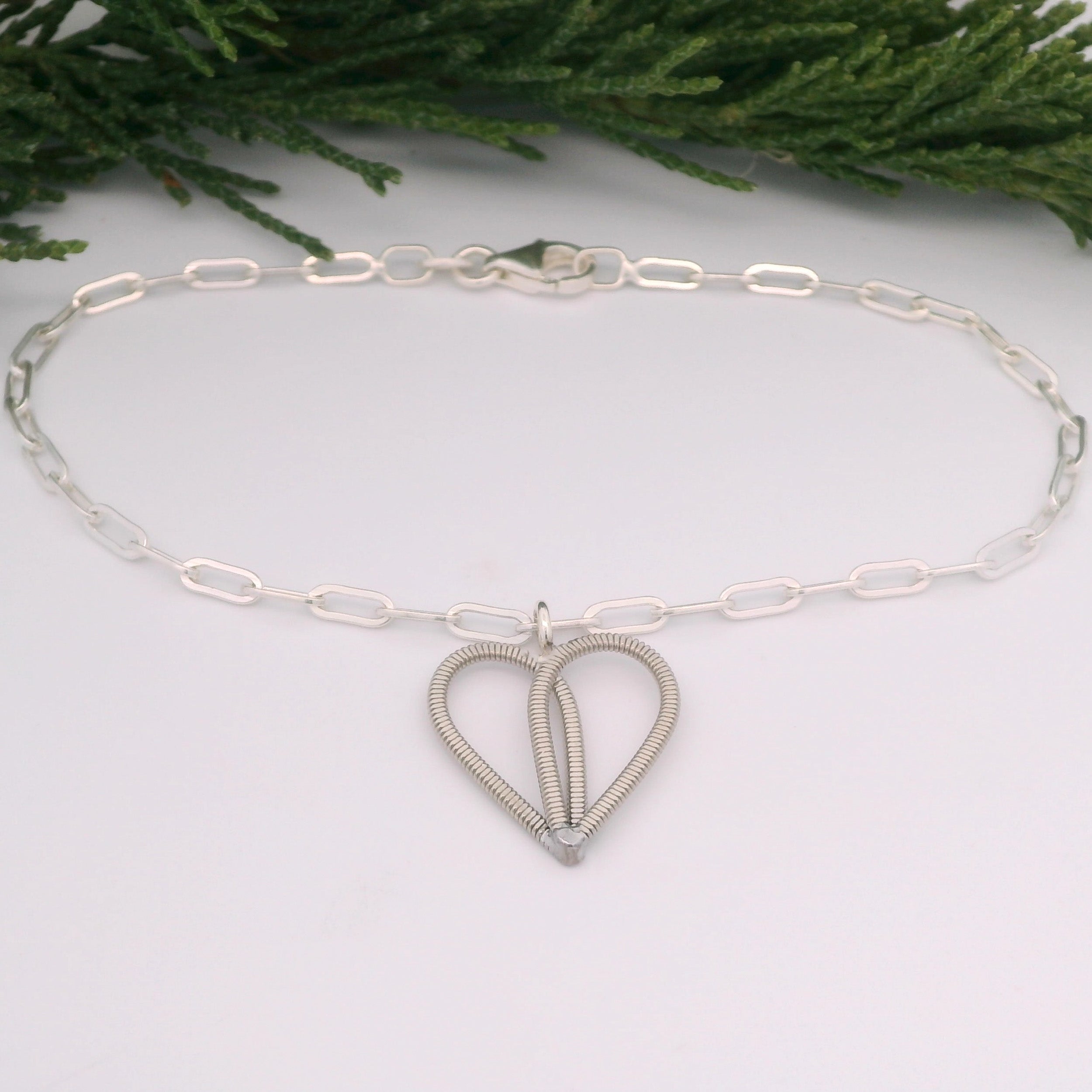 Silver paperclip chain bracelet with guitar string heart on white background with pine branch