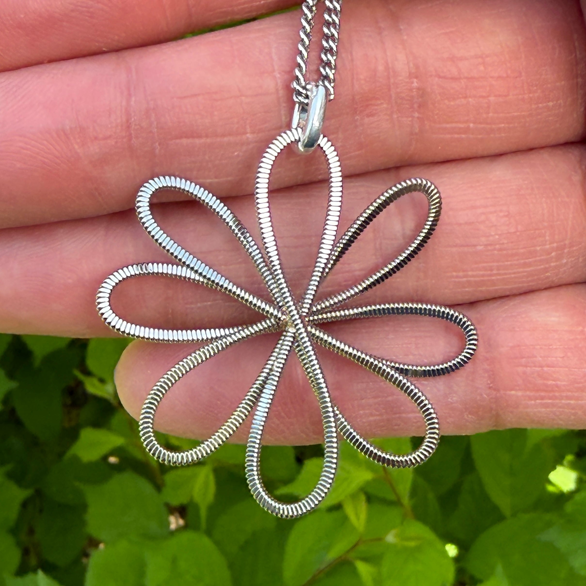 Silver floral guitar string pendant on a chain held between fingers with a green leafy background