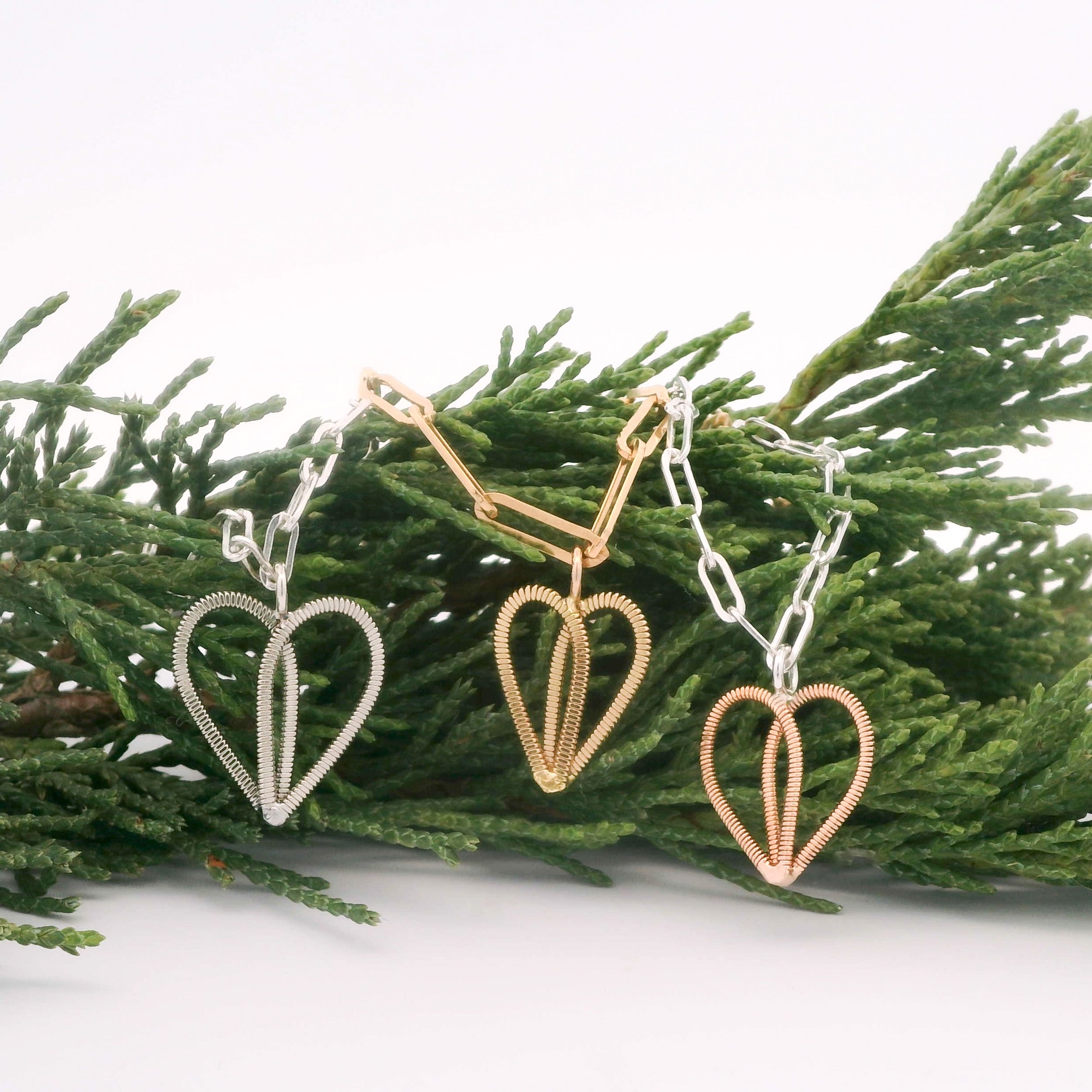 Gold guitar string heart on gold paperclip chain  and silver heart on silver paperclip chain and bronze heart on silver paperclip chain with white background and pine branch