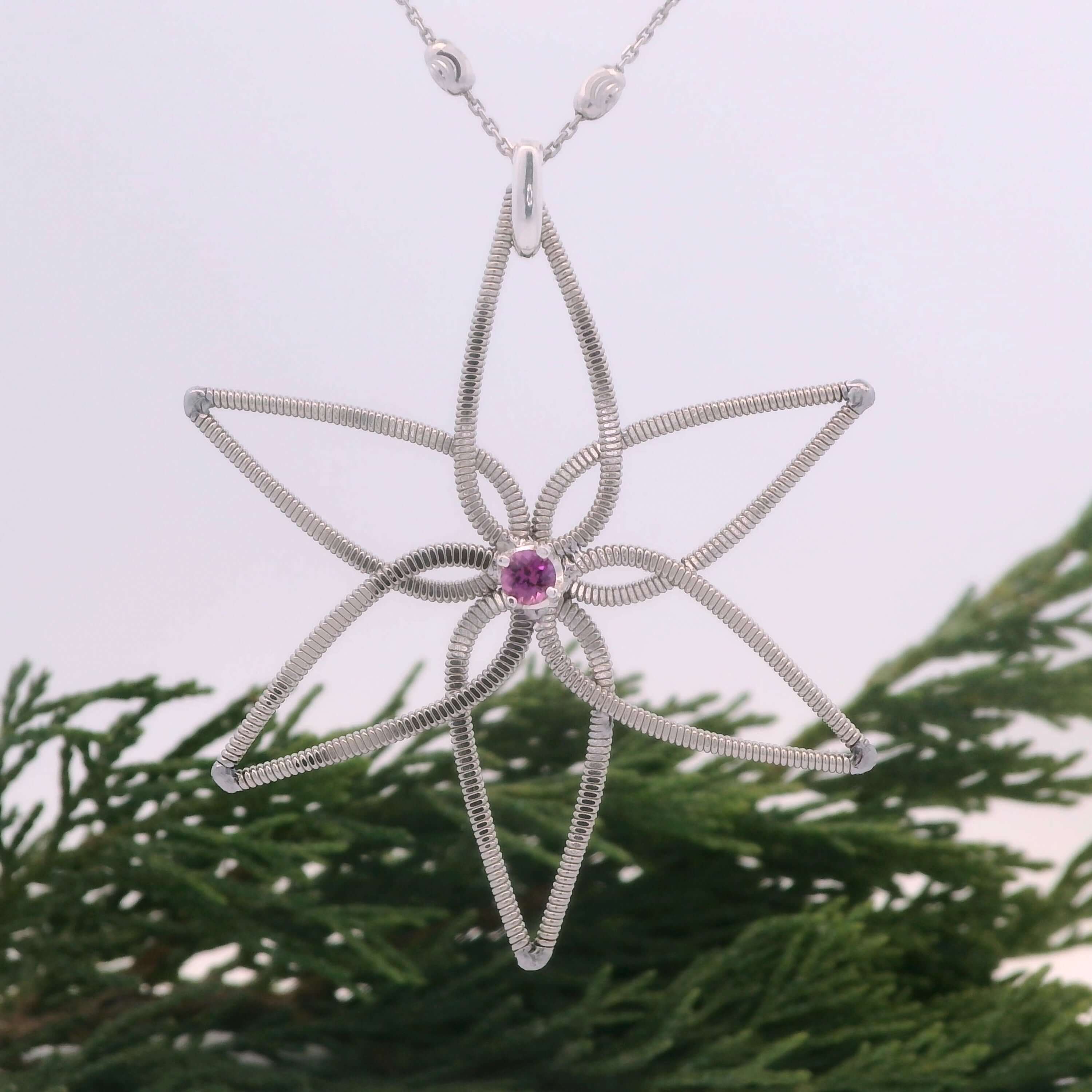 Electric guitar string flower pendant with a pink gemstone on a chain, set against a green leafy background.