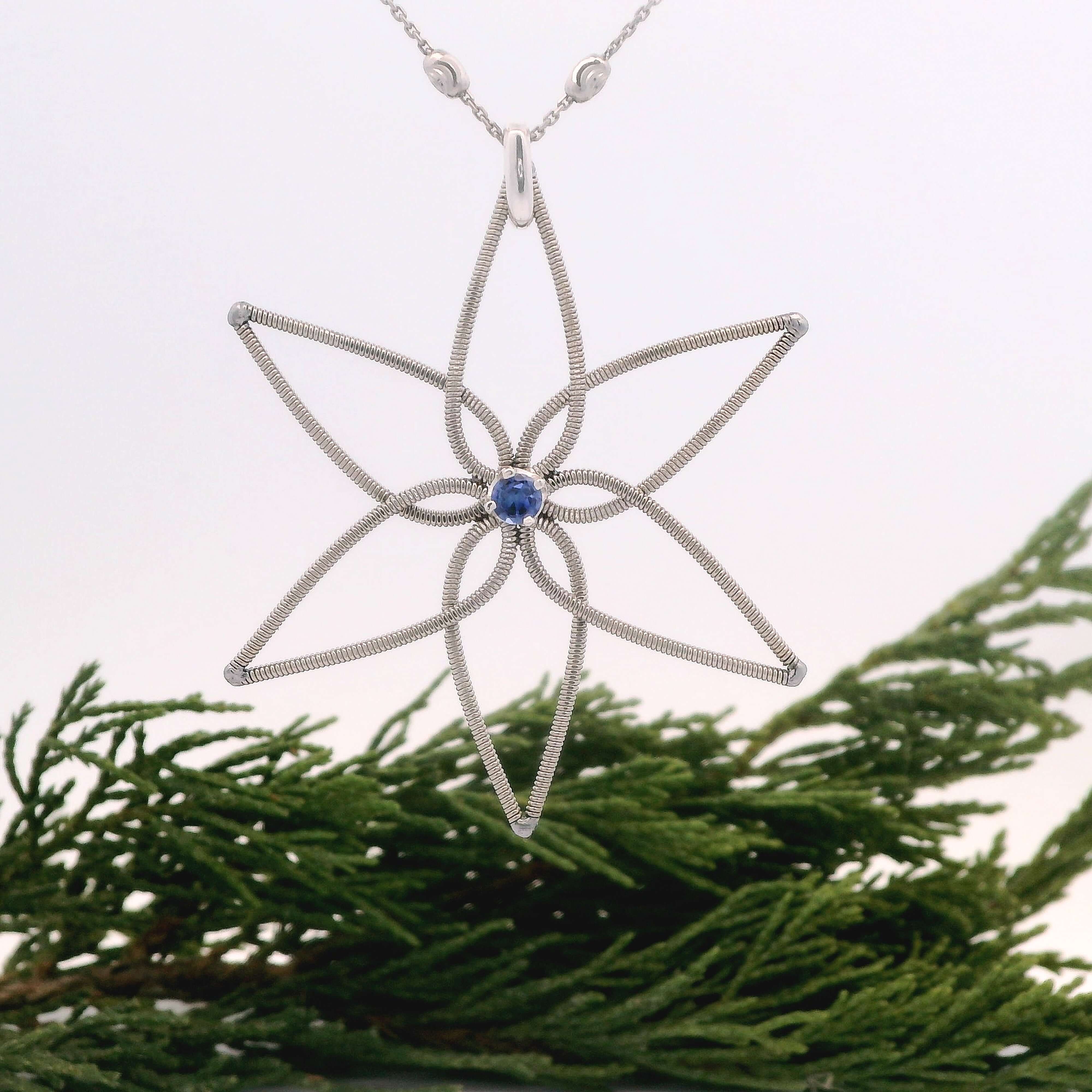 Electric guitar string flower pendant with a blue gemstone on a chain, set against greenery.