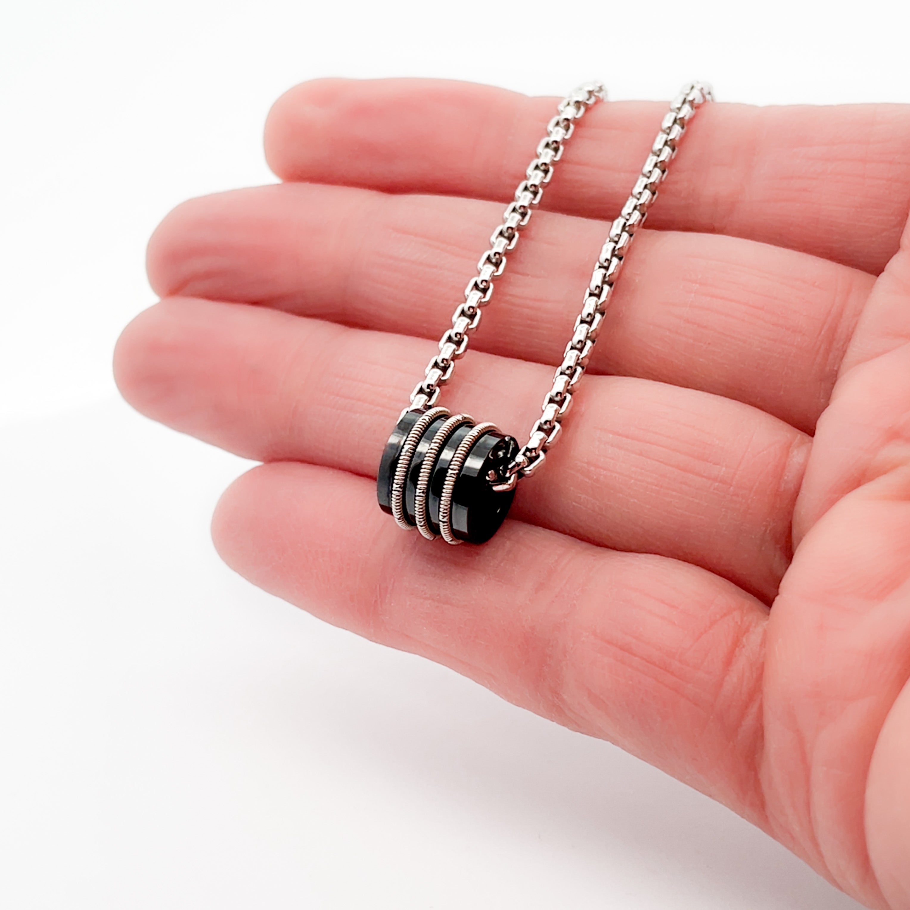 Silver necklace with black bead and inlaid guitar string on hand with white background.