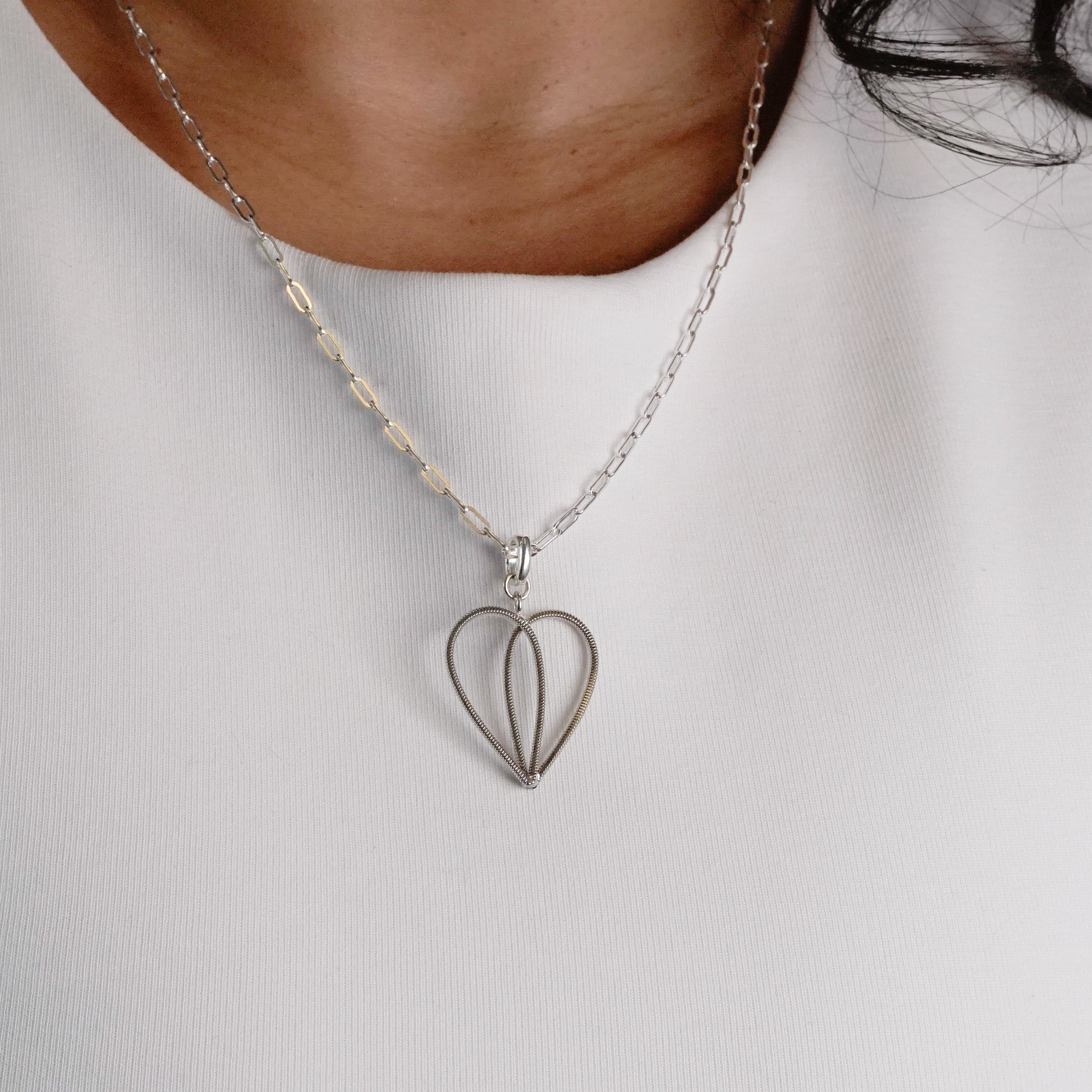 Silver heart-shaped guitar string pendant necklace on a person wearing a white shirt.