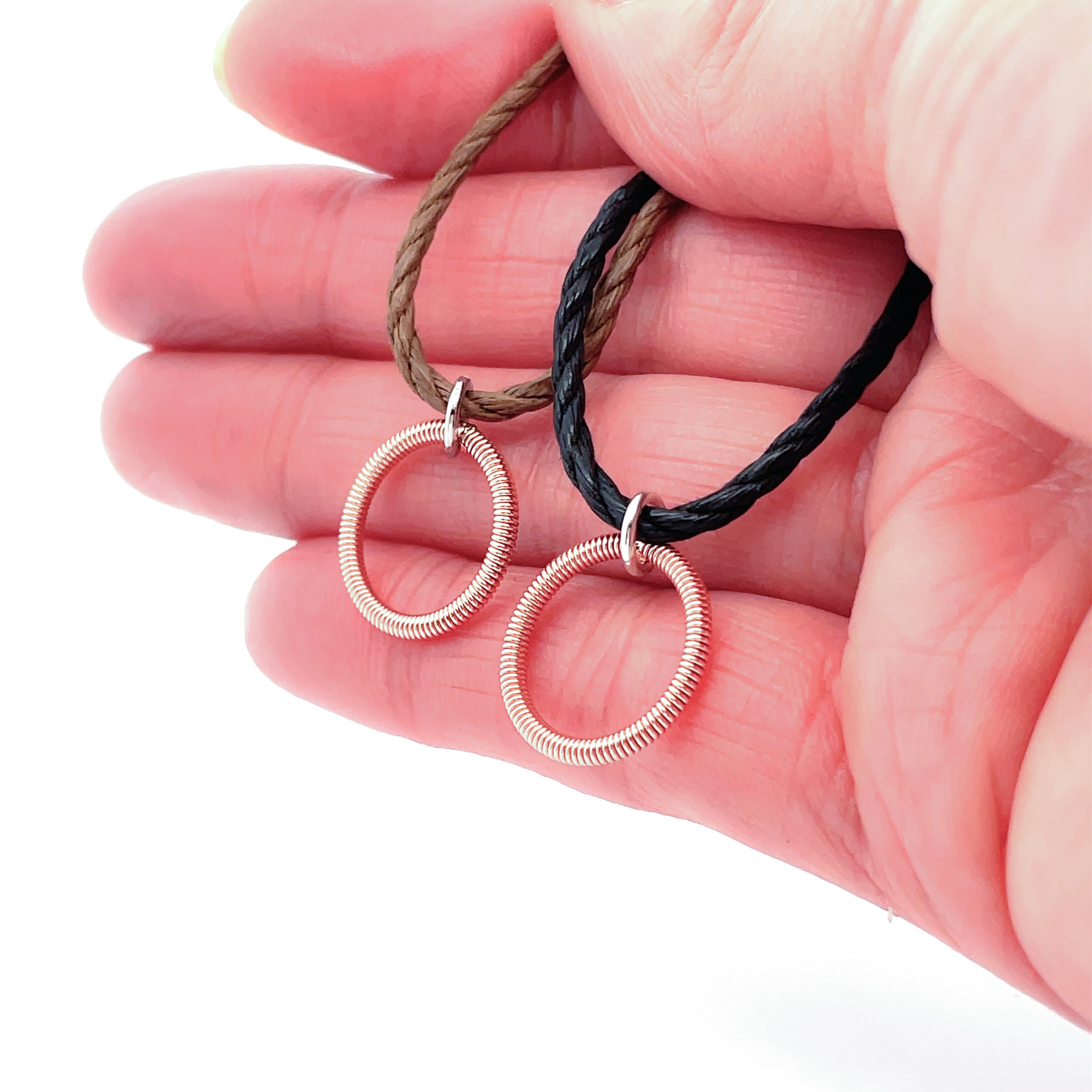 2 piano string circle pendants on black and brown cords in hand on white background